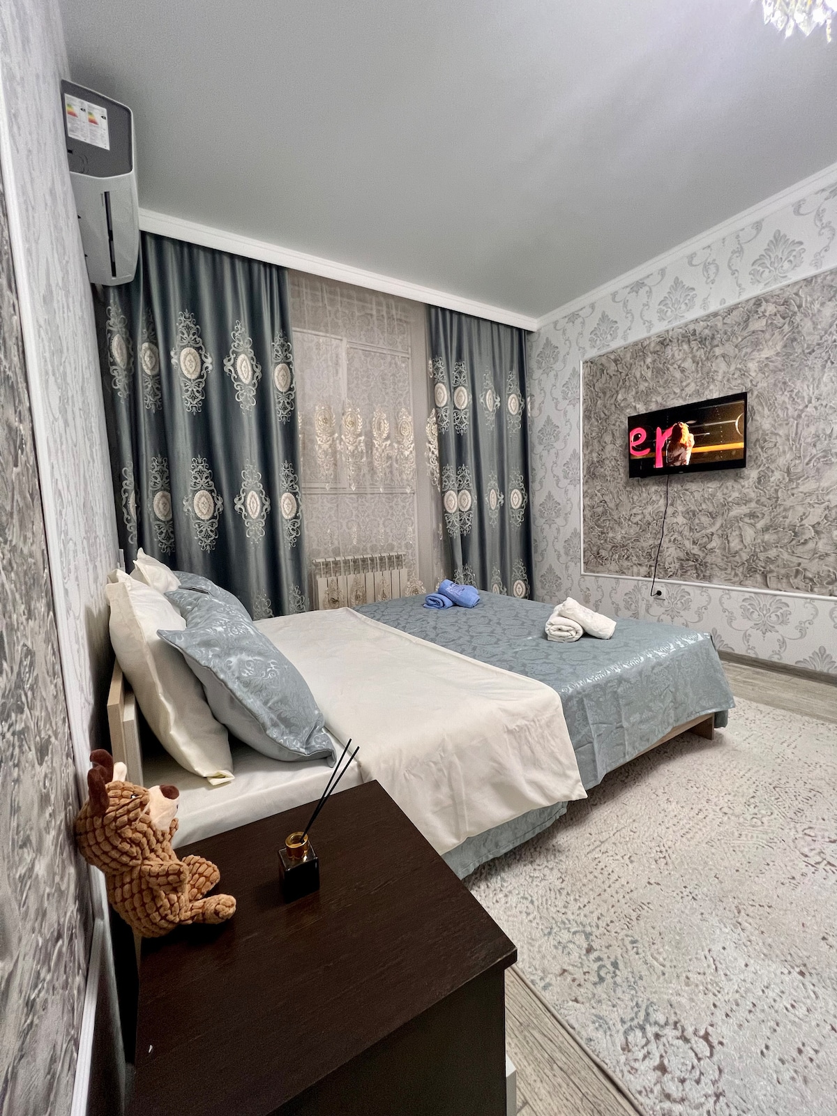 A well-decorated bedroom is highlighted by patterned wallpaper and soft curtains. A spacious bed is dressed with light-colored linens and towels are neatly arranged. A small wooden table with a teddy bear and an air conditioning unit adds comfort. A television is mounted on the wall.