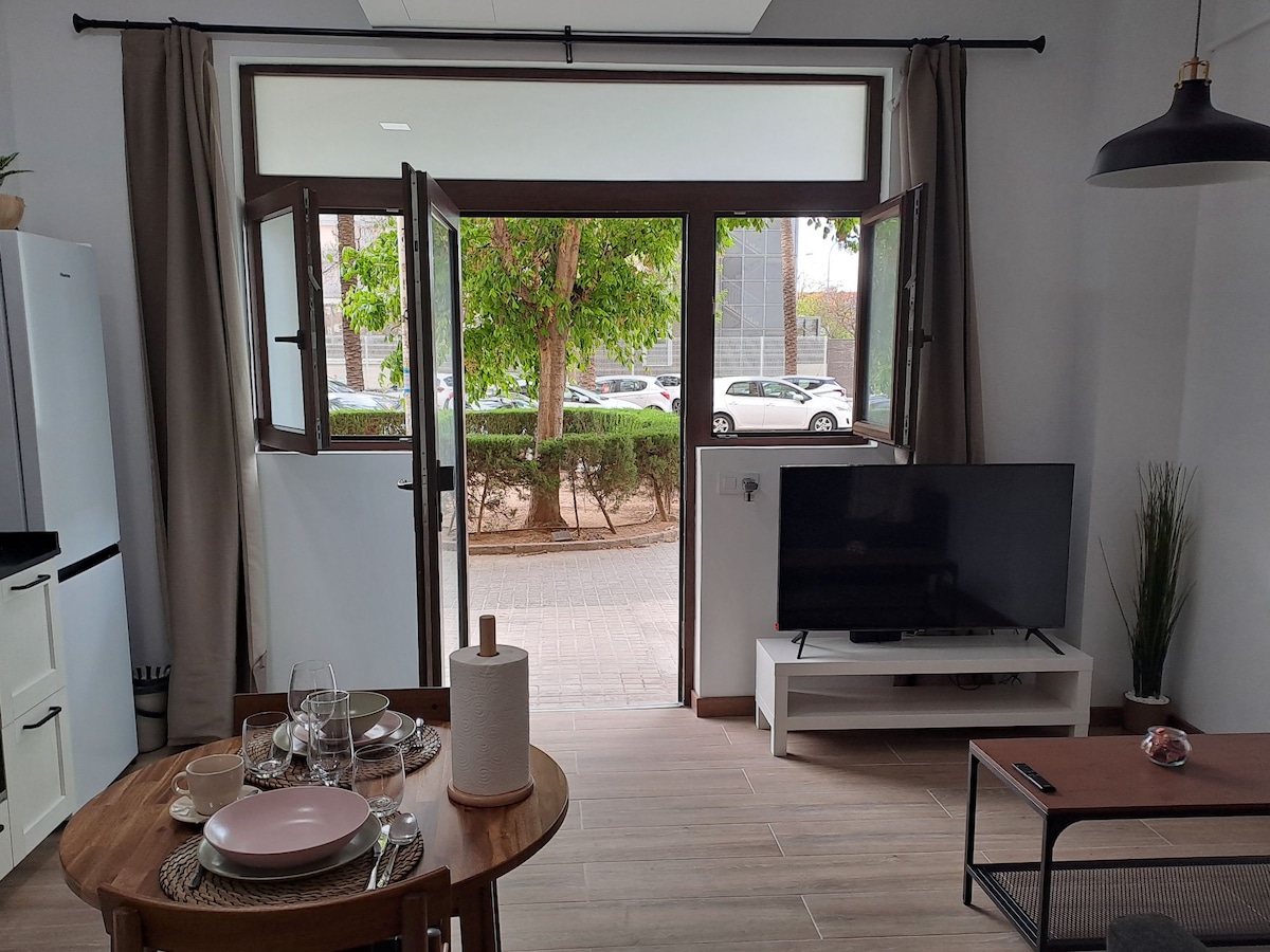 A bright living space is showcased with large windows providing views of greenery outside. The room features a dining table set for two, a comfortable seating area with a TV on a white console, and natural light streaming in through the open windows.