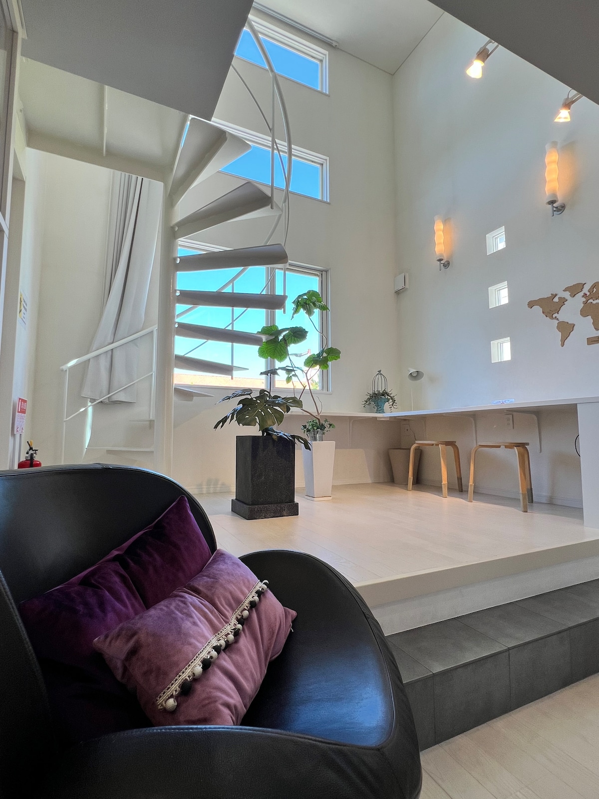 A spiral staircase leads to a sunlit space featuring large windows. A cozy black chair with purple pillows sits in the foreground, while a small plant is placed on a pedestal nearby. Natural light illuminates minimalist decor, enhancing the airy atmosphere.
