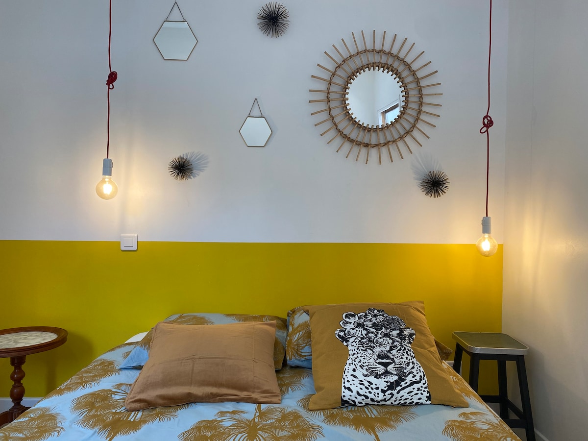 A cozy sleeping area features a brightly colored accent wall, layered with soft bedding and decorative pillows. Two pendant lights hang above the bed, providing illumination. Decorative mirrors and wall art add texture and interest to the space.