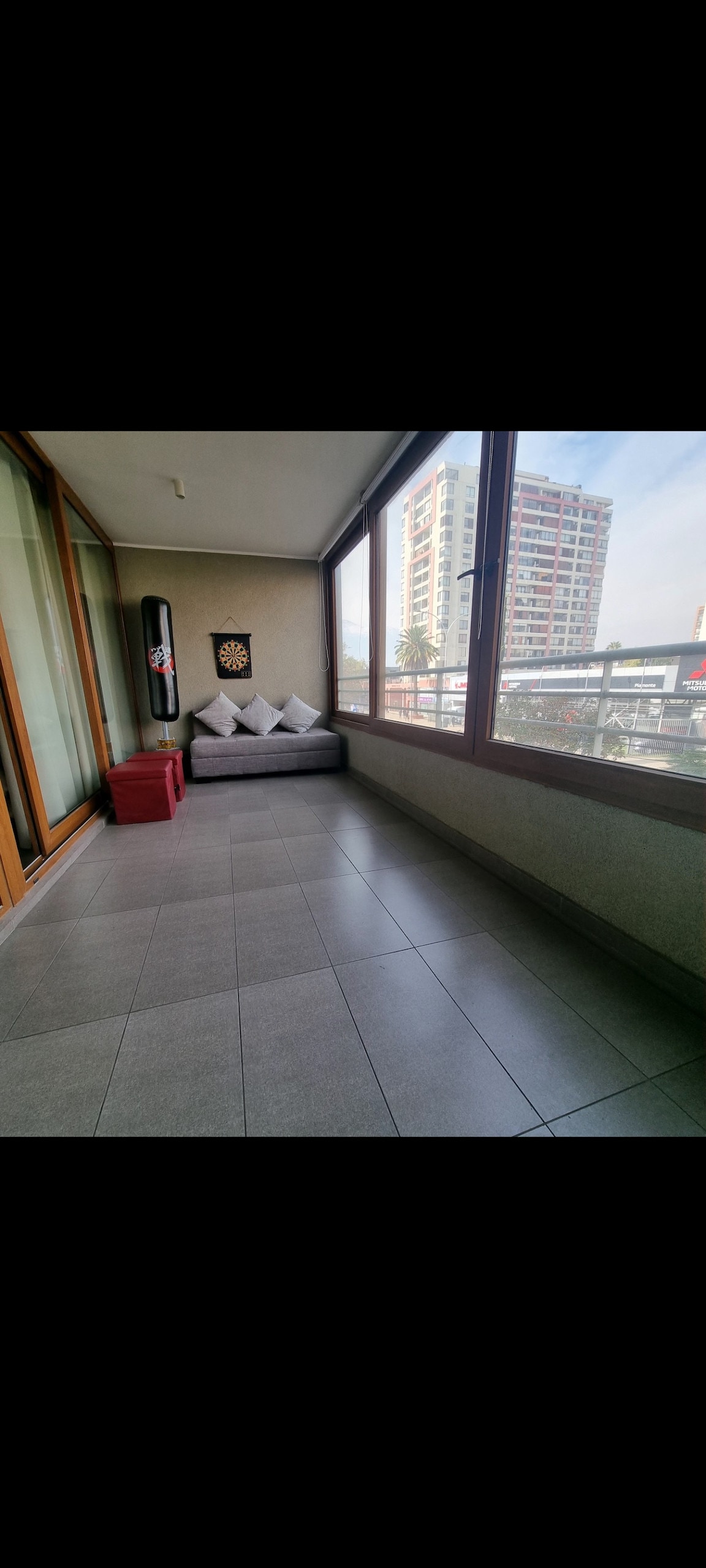 A bright terrace is shown, featuring large glass windows that allow natural light to flood the space. A gray sofa with decorative cushions is positioned against the wall, complemented by a small red storage box. The smooth tiled floor reflects the urban surroundings visible outside.