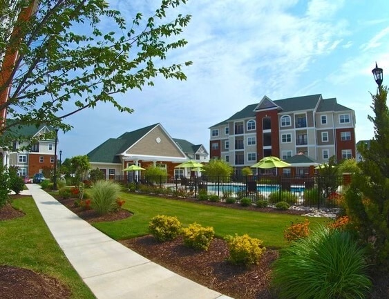 A landscaped pathway winds through lush greenery, leading to a vibrant pool area. The multi-story building features a mixture of grey and red facades, while chairs are arranged under bright umbrellas. Trees and flowering plants enhance the inviting outdoor space.