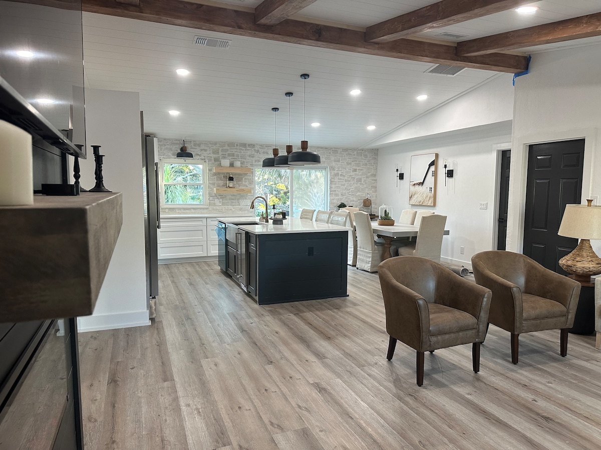 An open-concept living area features a kitchen island with seating, surrounded by modern cabinetry and light-colored walls. Natural light enters through windows, illuminating the room. Two brown armchairs are situated near a fireplace, while a dining area with stylish seating is visible in the background.