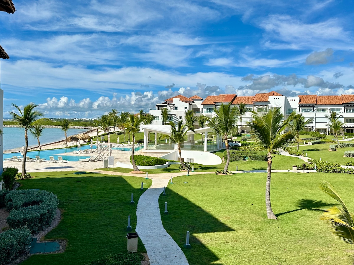 A tropical landscape showcases a well-maintained garden, featuring palm trees and pathways leading to the infinity pool by the ocean. Residential buildings reflect a modern architectural style. The sky is blue with scattered clouds, creating a serene atmosphere.