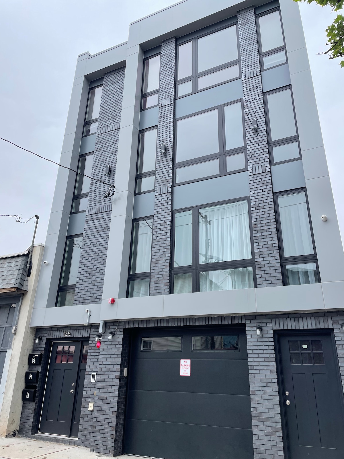 A modern three-story building is displayed, featuring large windows with a black frame and a sleek grey exterior. The façade includes a combination of brick and smooth surfaces, while the entrance is marked by a dark garage door and adjacent doors for access.