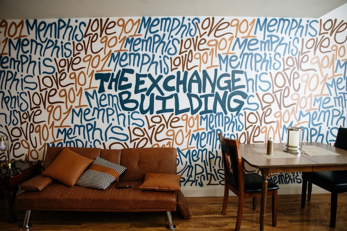 A vibrant wall mural showcases the name 'The Exchange Building' prominently alongside the word 'Memphis' in various styles. A modern couch with brown cushions and a dining table with black chairs are positioned on a polished wooden floor, creating a welcoming living space.