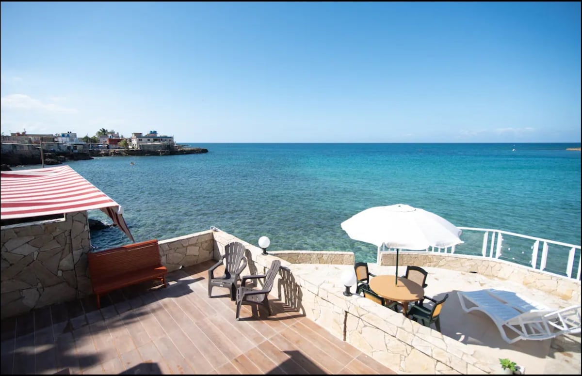 A spacious terrace overlooks the tranquil bay, featuring a round table and four chairs shaded by a large umbrella. The area is surrounded by natural stone walls, with the calm turquoise waters visible just beyond the edge, offering a serene atmosphere by the sea.