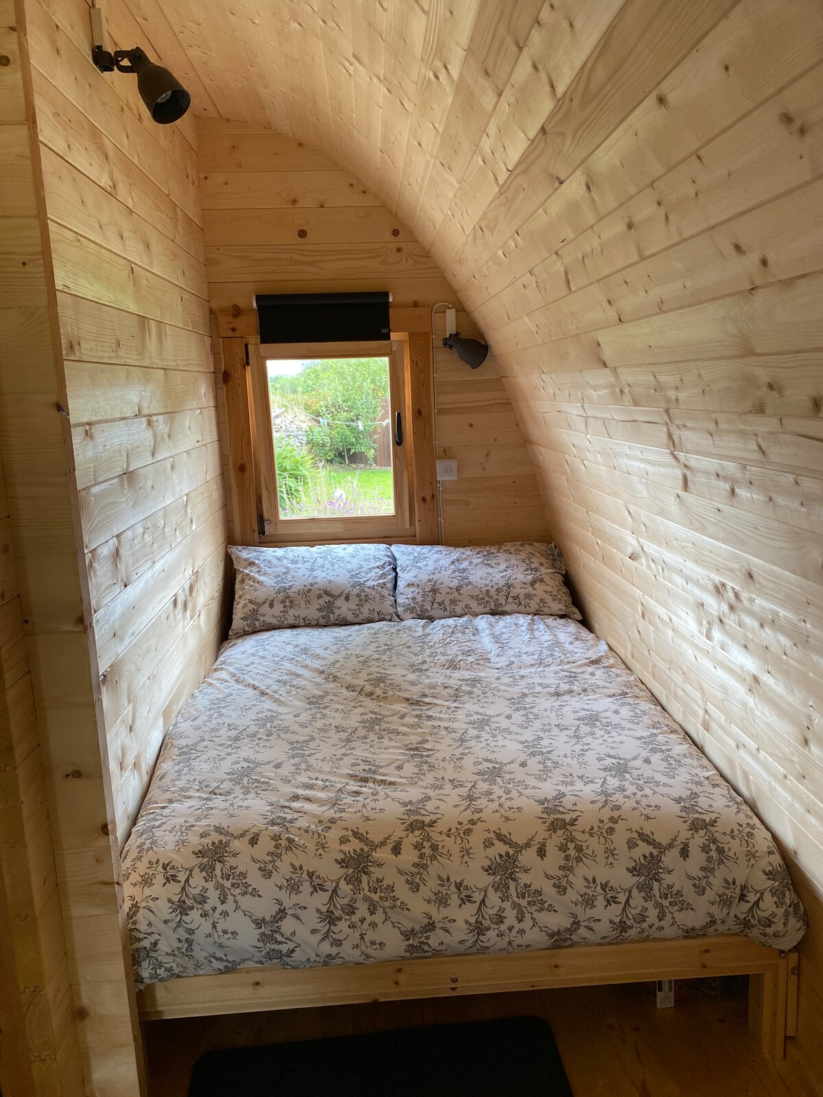 A cozy sleeping area features a spacious double bed adorned with floral-patterned bedding. Warm wooden walls create a natural ambiance. A small window allows natural light to enter, revealing green outdoor surroundings. Simple decor emphasizes a tranquil environment.