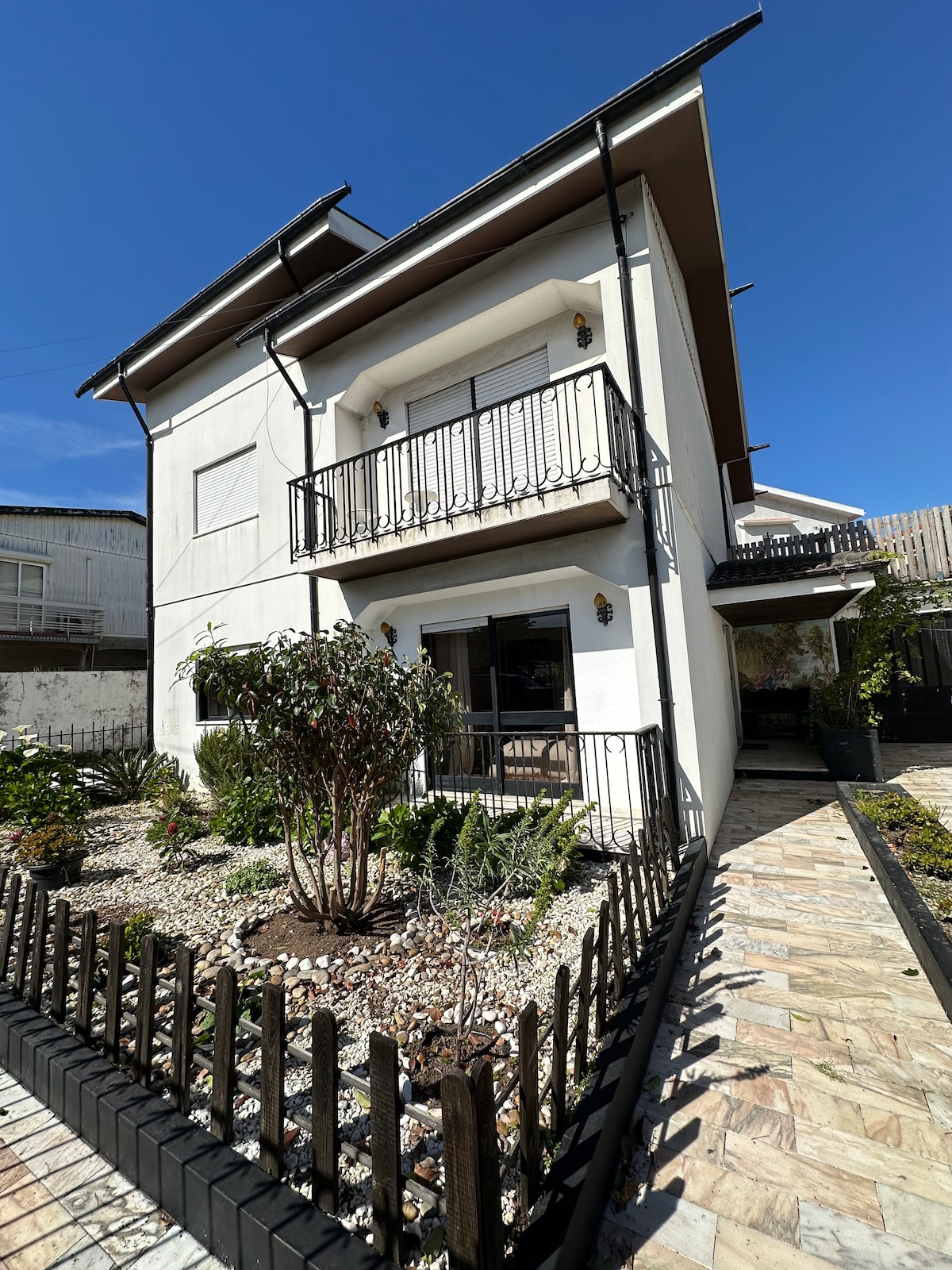 The exterior of a three-story house is shown, featuring a balcony on the second floor. A small garden with shrubs and decorative stones lines the front, while a paved pathway leads to the entrance under clear blue skies.