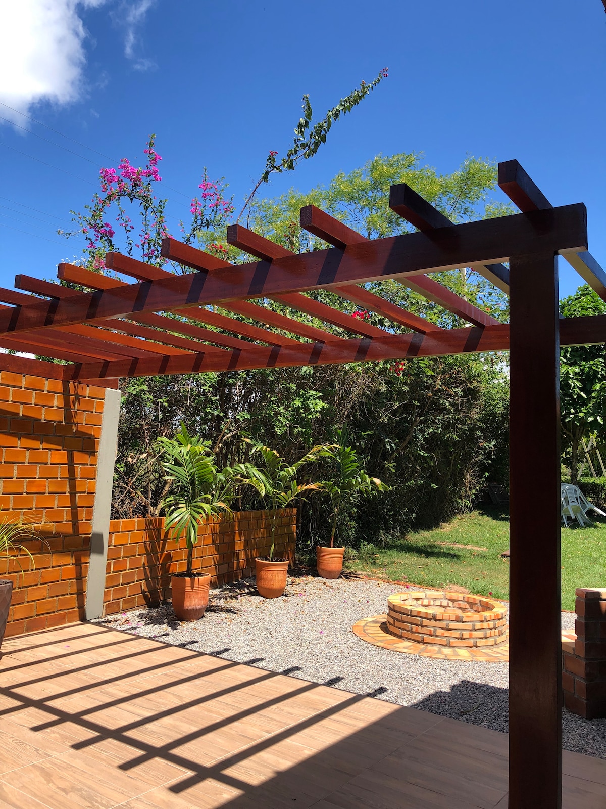 A wooden pergola stands against a clear blue sky, providing partial shade. Potted plants are arranged around a gravel area, leading to a circular stone fire pit at the center. Lush greenery surrounds the space, enhancing the outdoor environment.