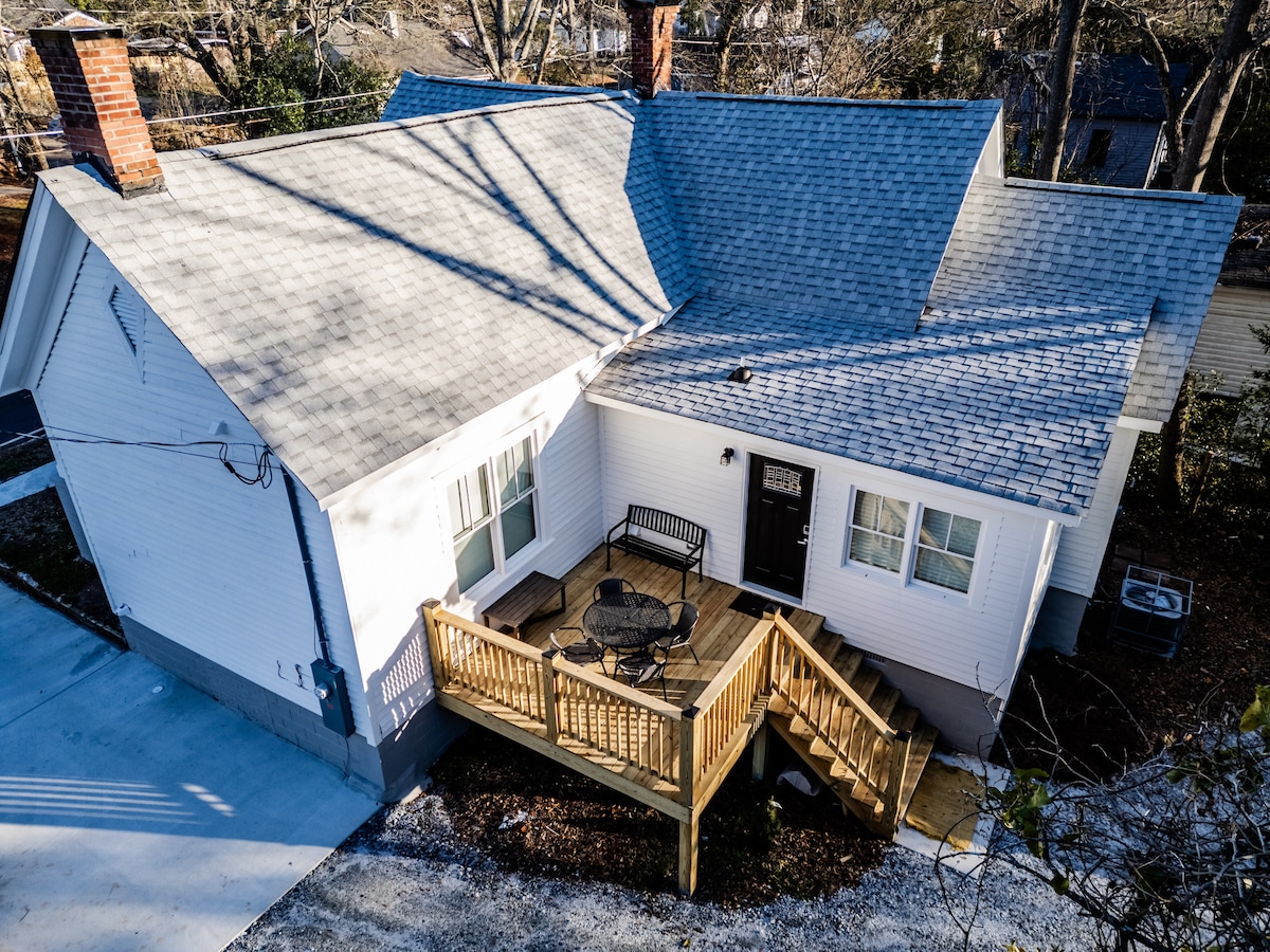 An aerial view of the cottage is shown, highlighting the front deck surrounded by a wooden railing. The exterior features light-colored siding and a grey roof, with a small seating area set up for outdoor relaxation. Nearby, a paved driveway leads up to the home.