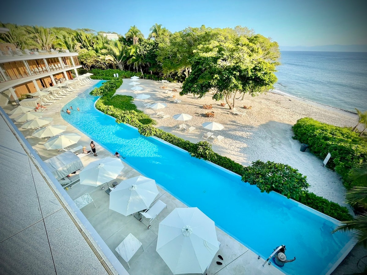 A winding infinity pool stretches alongside a sandy beach, flanked by lounge chairs and umbrellas. Lush greenery and palm trees provide shade, while the expansive ocean reflects the clear sky. The tranquil setting invites relaxation and enjoyment of the natural surroundings.