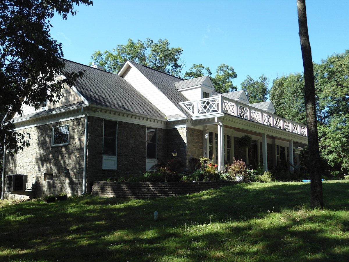 A spacious home is featured, showcasing a blend of stone and siding. The front porch with white railing invites relaxation, surrounded by lush greenery. Large windows enhance the connection to nature, highlighting the serene atmosphere of the surrounding property.