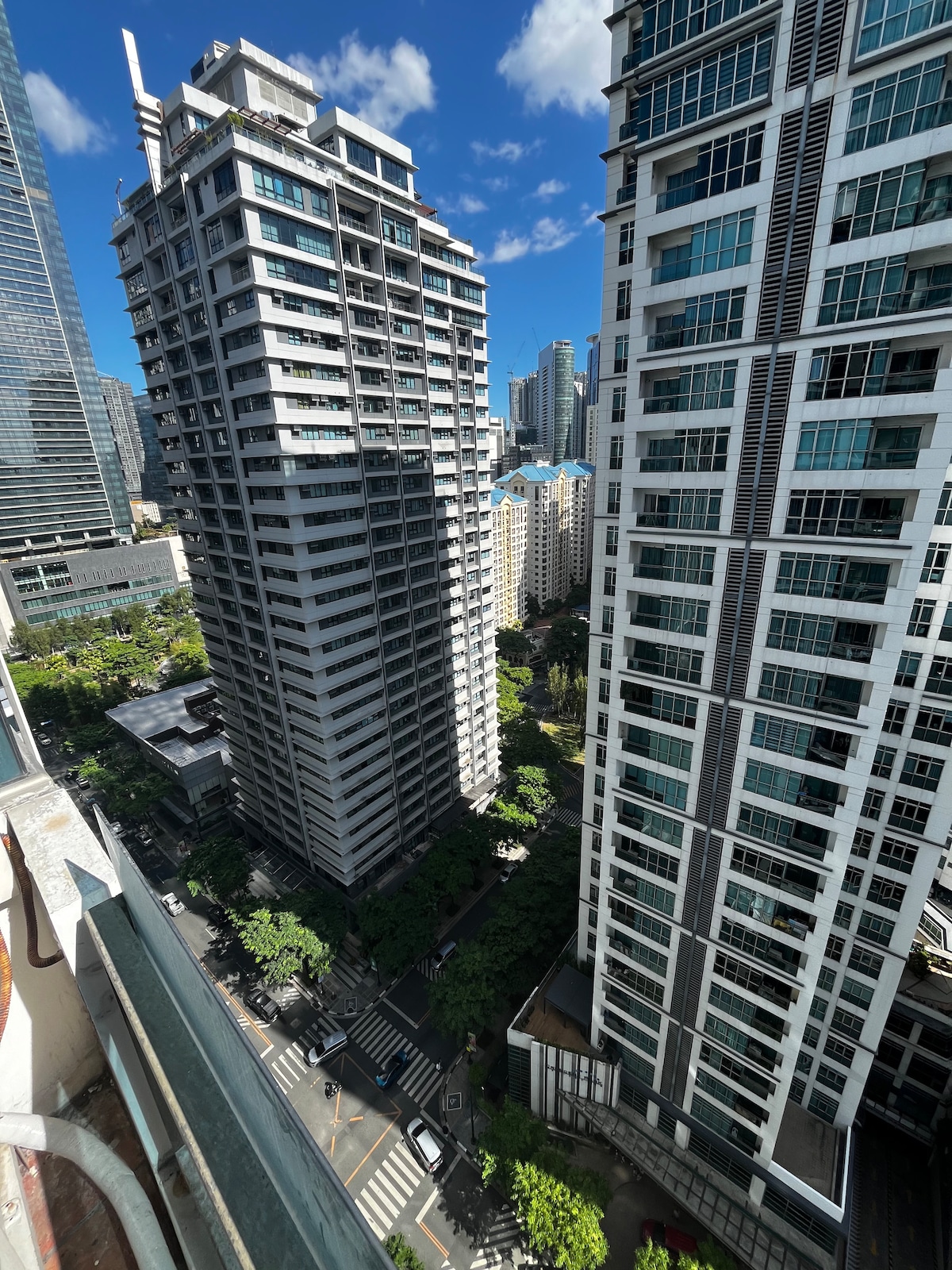 The view captures a vibrant urban landscape featuring tall residential buildings. Lush greenery is visible at street level, contrasting with the modern architecture. A clear blue sky provides natural lighting, enhancing the overall impression of the bustling environment below.