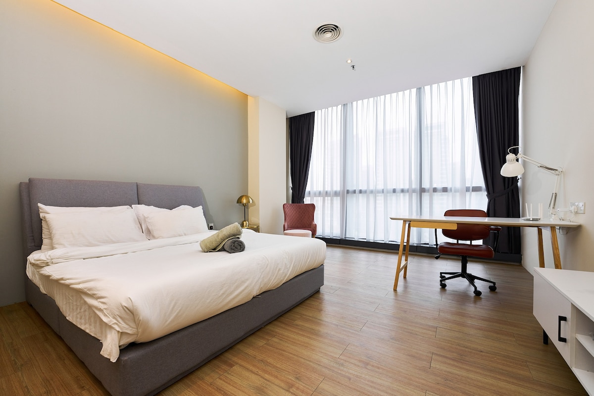 A spacious sleeping area features a large bed with crisp linens and neatly arranged pillows. A comfortable chair is positioned nearby, while a well-lit workspace is provided with a desk and an ergonomic chair. Large windows allow natural light to fill the room.