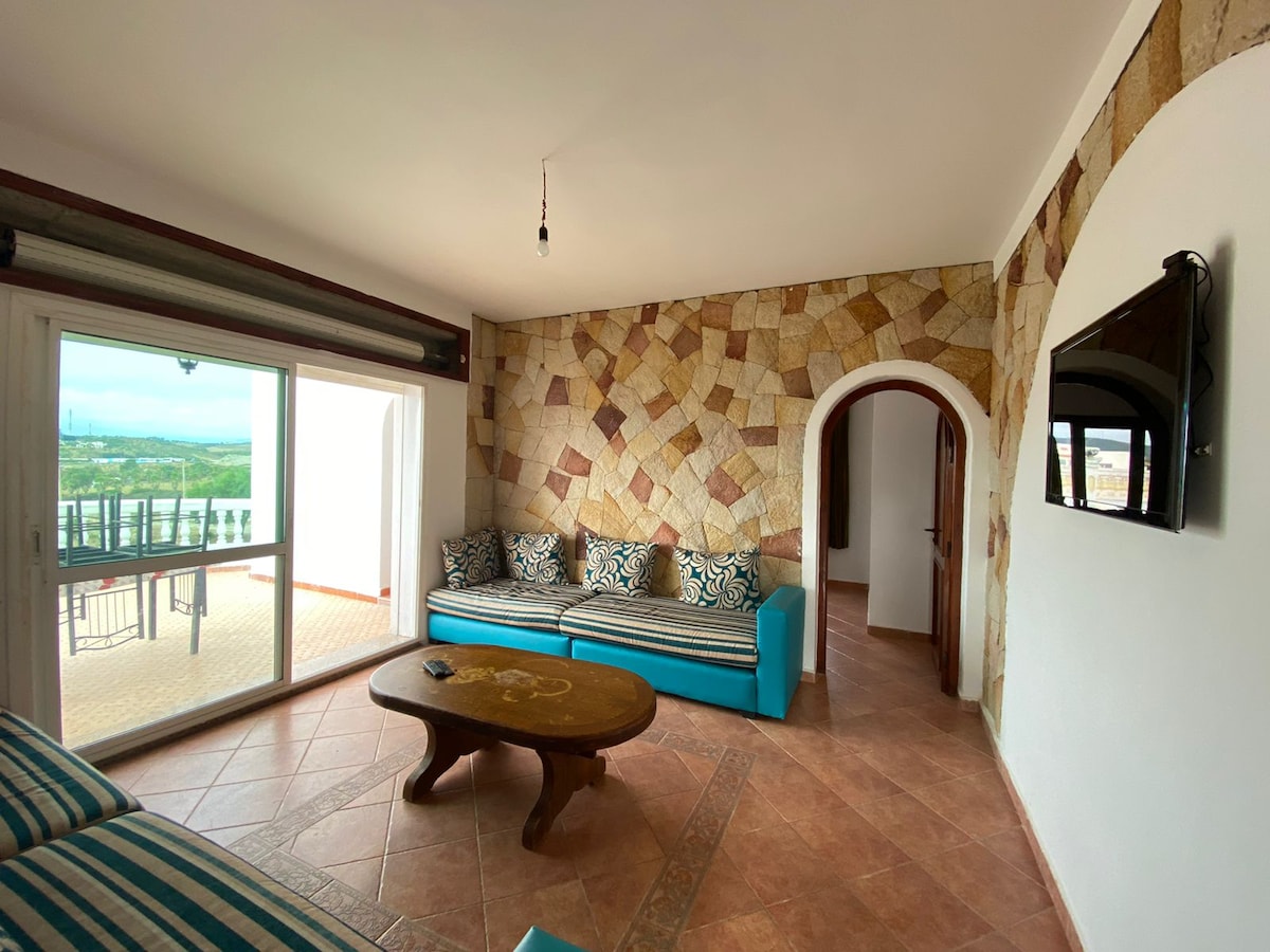 A comfortable sitting area is presented with a turquoise sofa adorned with patterned cushions. An organic stone wall adds texture, while a wooden coffee table sits in the center. A sliding door opens to a balcony, providing panoramic views of the surrounding landscape.