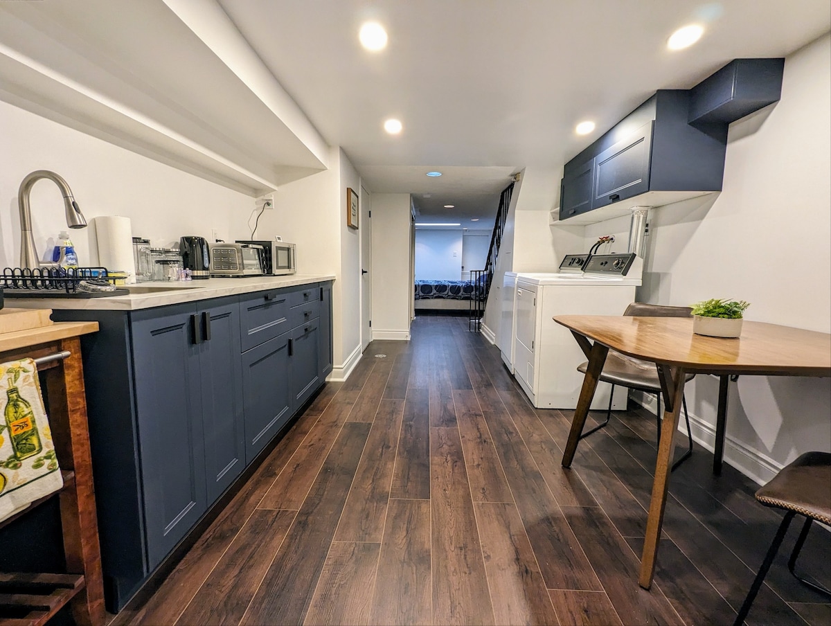 A narrow kitchen area features dark cabinetry and white appliances, including a washer and dryer. A small dining table with legs is positioned near the entrance, with warm lighting illuminating the space. The flooring is dark wood, enhancing the modern feel.