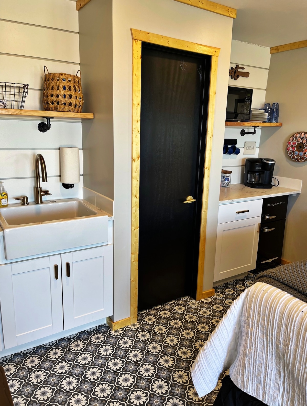 The kitchenette area includes a farmhouse-style sink with an elegant black cabinet door nearby. Stylish floating shelves hold decorative baskets and kitchen essentials. A coffee maker rests on the countertop next to a dark cabinet, while a patterned floor adds character to the space.