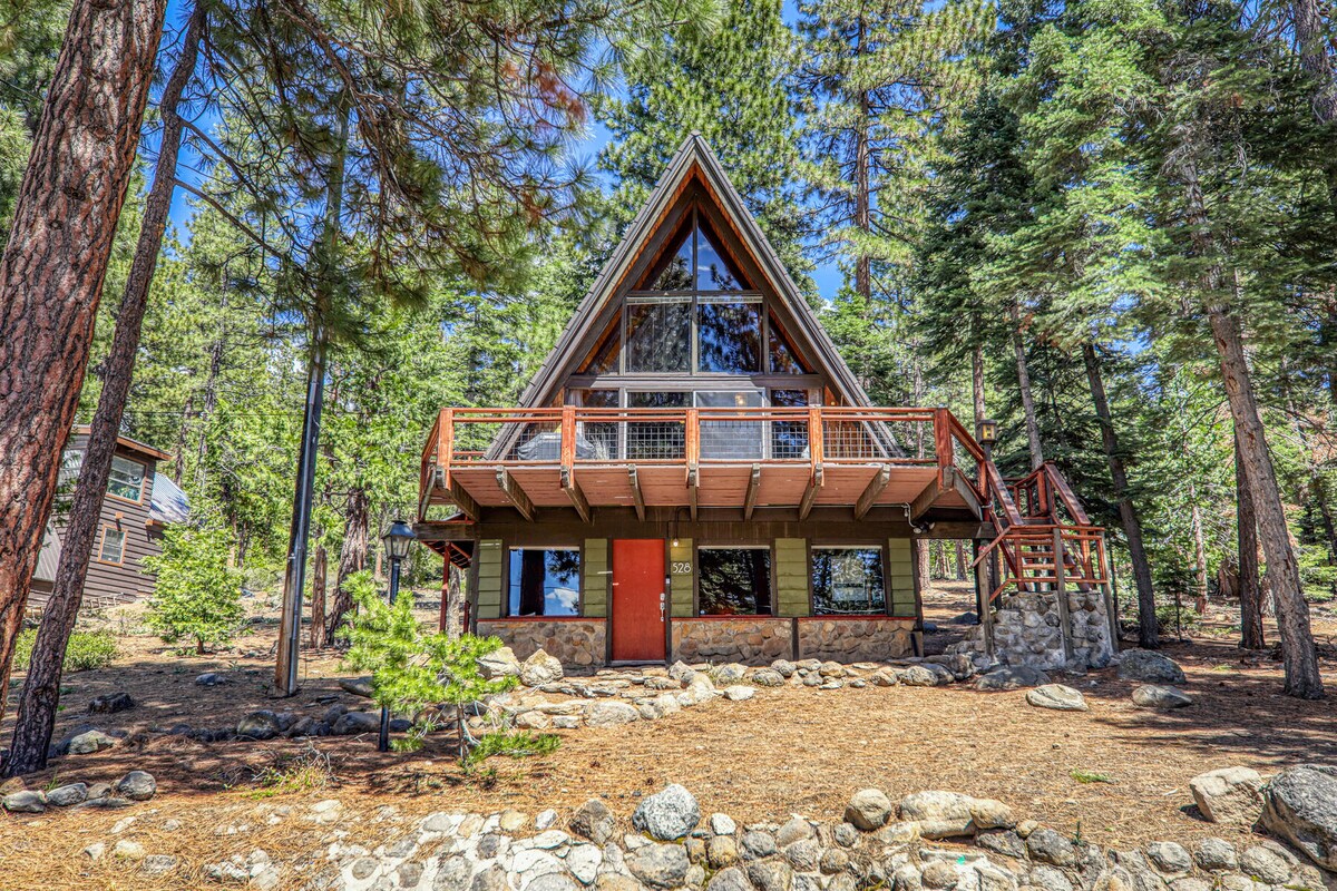 The A-Frame cabin showcases a distinctive triangular design with large front windows, allowing for natural light to fill the interior. The deck extends from the main level, offering outdoor access. Surrounded by pine trees and a natural landscape, the home blends harmoniously with its environment.