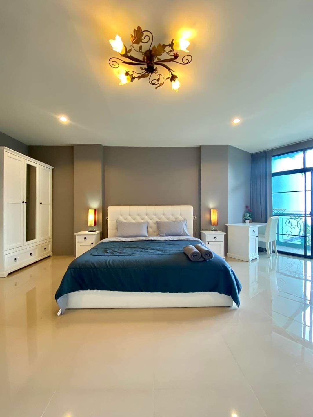 The spacious bedroom features a large bed with a dark blue blanket and matching towels. A white wardrobe is positioned beside the bed, and a glass door leads to a balcony. Soft lighting enhances the room's calm ambiance, complemented by a decorative chandelier.