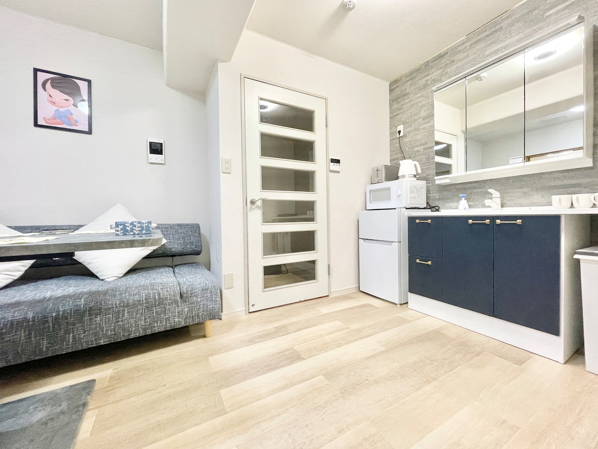 A bright room features a textured gray sofa adorned with decorative pillows, positioned beside a sleek, modern kitchenette. A mirrored cabinet reflects light, with a compact refrigerator and microwave visible. A glass-paned door leads to another space, enhancing the open feel of the area.
