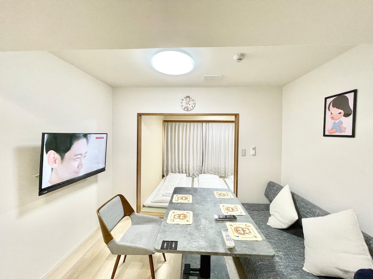 A modern living space features a cozy dining area with a table and upholstered chairs. A wall-mounted television is visible, alongside a framed artwork. Soft, natural light enters through a circular ceiling fixture, while a partition leads to a sleeping area with neatly made beds.