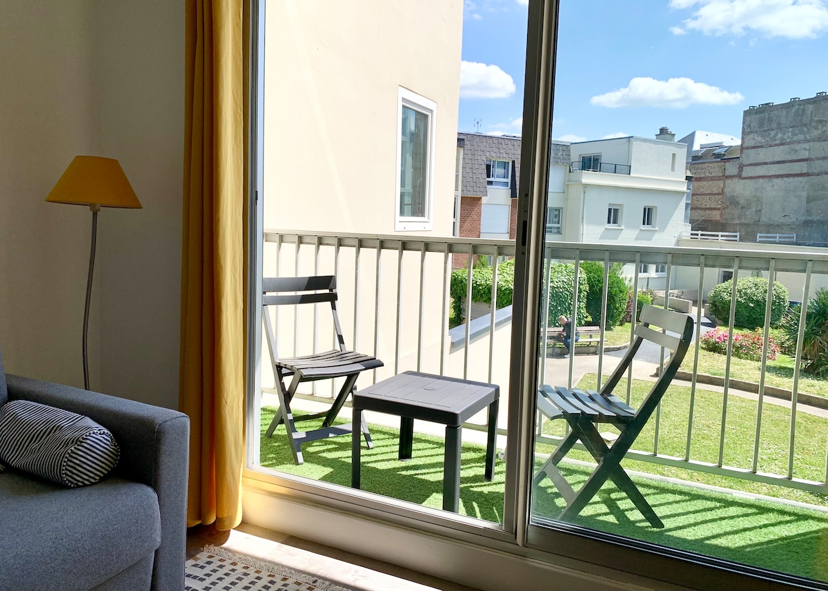 The image displays a balcony with two folding chairs and a small table, overlooking a green courtyard. Natural light floods the space through large glass doors, framed by soft curtains, creating a serene ambiance in the living area.