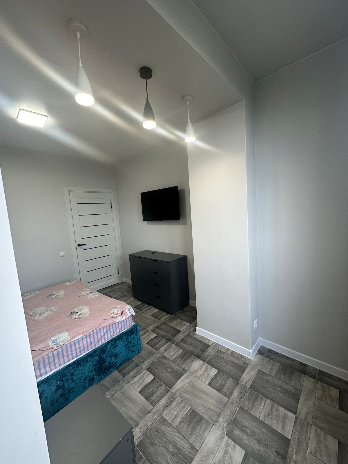 A well-lit bedroom features a spacious bed covered with a soft pink blanket and whimsical patterns. A dark dresser stands against the wall, and a television is mounted above, providing entertainment options. The room is complemented by modern pendant lighting and contemporary tile flooring.