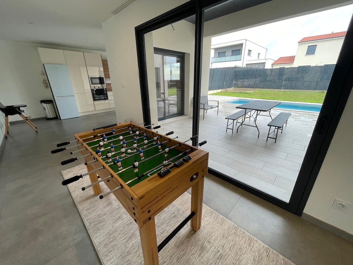 A foosball table is positioned in a light-filled area, with large glass doors providing a view of the grassy outdoor space and pool. A dining table is visible on the patio, offering seating options for relaxation and enjoyment.