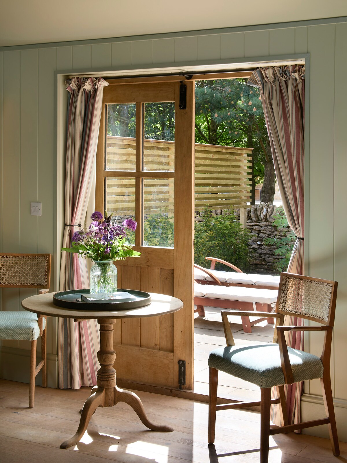 A wooden door with glass panes opens to a private terrace, framed by light curtains. A small round table sits at the forefront, accompanied by two chairs. A vase of fresh flowers adds a touch of color, while sunlit greenery is visible beyond the door.
