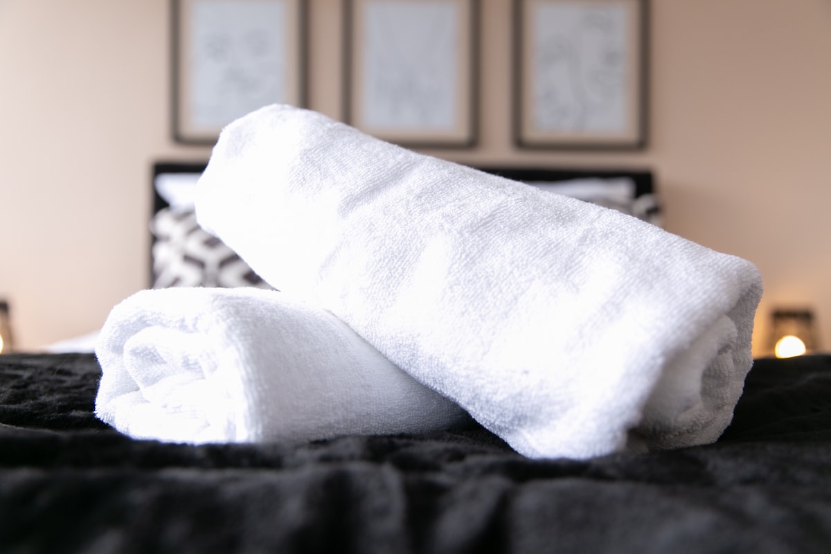Two neatly rolled white towels are placed on a dark bedding surface. A blurred background reveals a stylish bed with patterned pillows and framed art on the wall, contributing to a clean and organized environment.