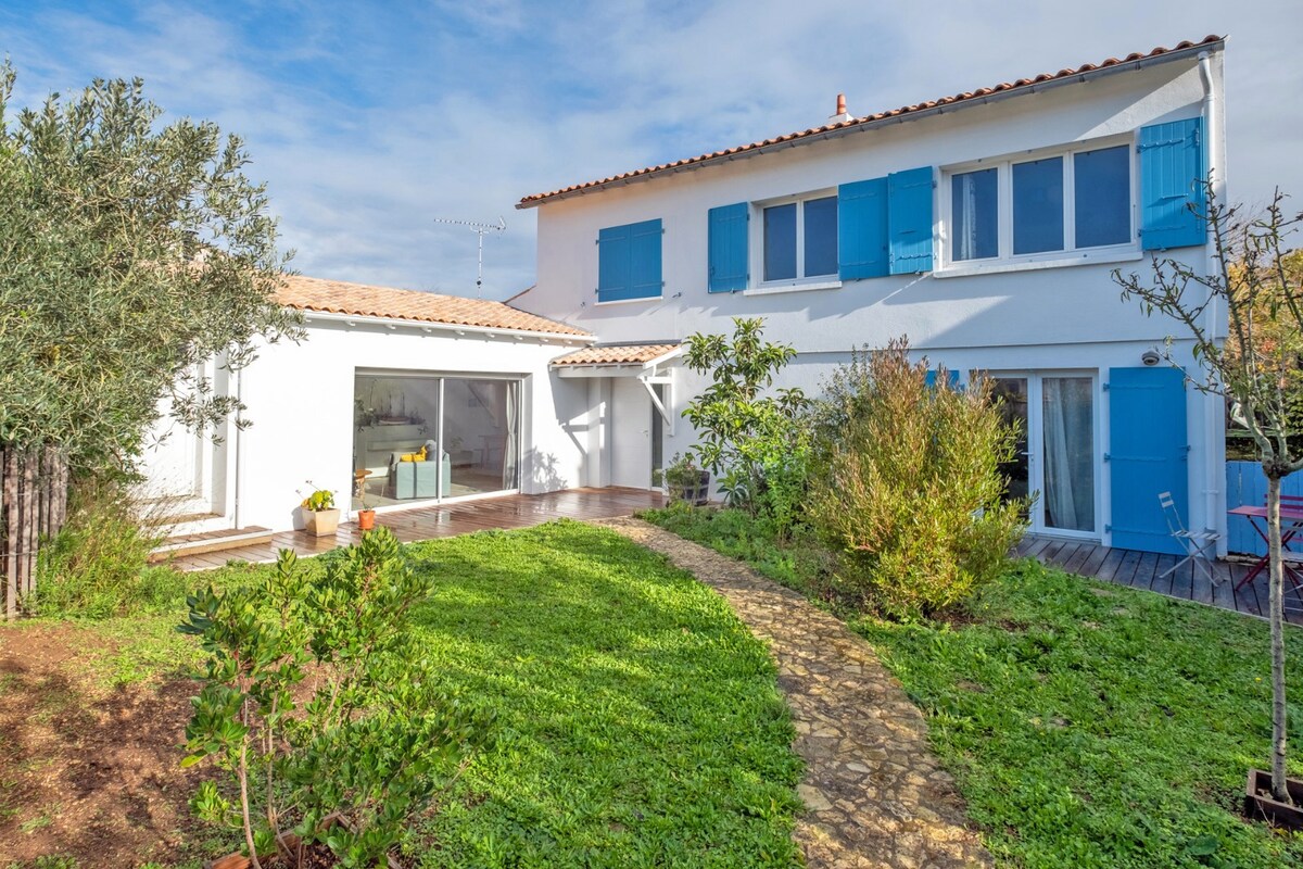 The exterior of a white two-story house is showcased, featuring blue shutters and a tiled roof. A wooden deck is visible, leading to a landscaped garden with a stone pathway and lush greenery. The background is enhanced by various plants and trees surrounding the property.
