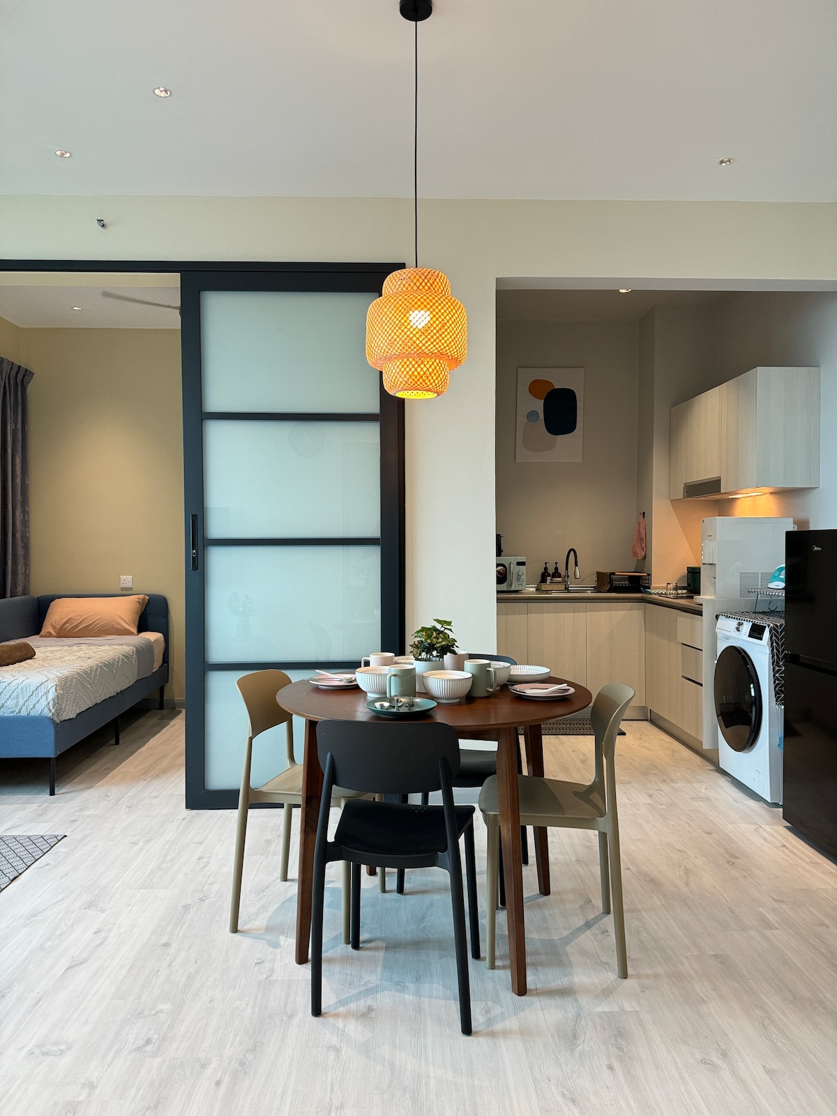 A dining area is presented with a round table surrounded by four chairs, set for a meal with dishes and cutlery. A modern pendant light hangs from the ceiling, illuminating the space. The kitchen and living area are visible in the background, enhancing the openness.