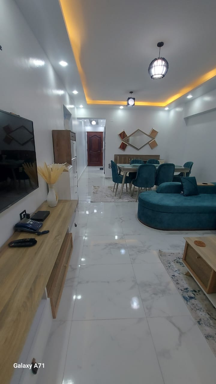 A modern living space features polished marble flooring with a light, airy feel. A sleek television is mounted on the wall opposite a stylish sofa bed and a dining table surrounded by upholstered chairs. Soft lighting complements the contemporary decor.