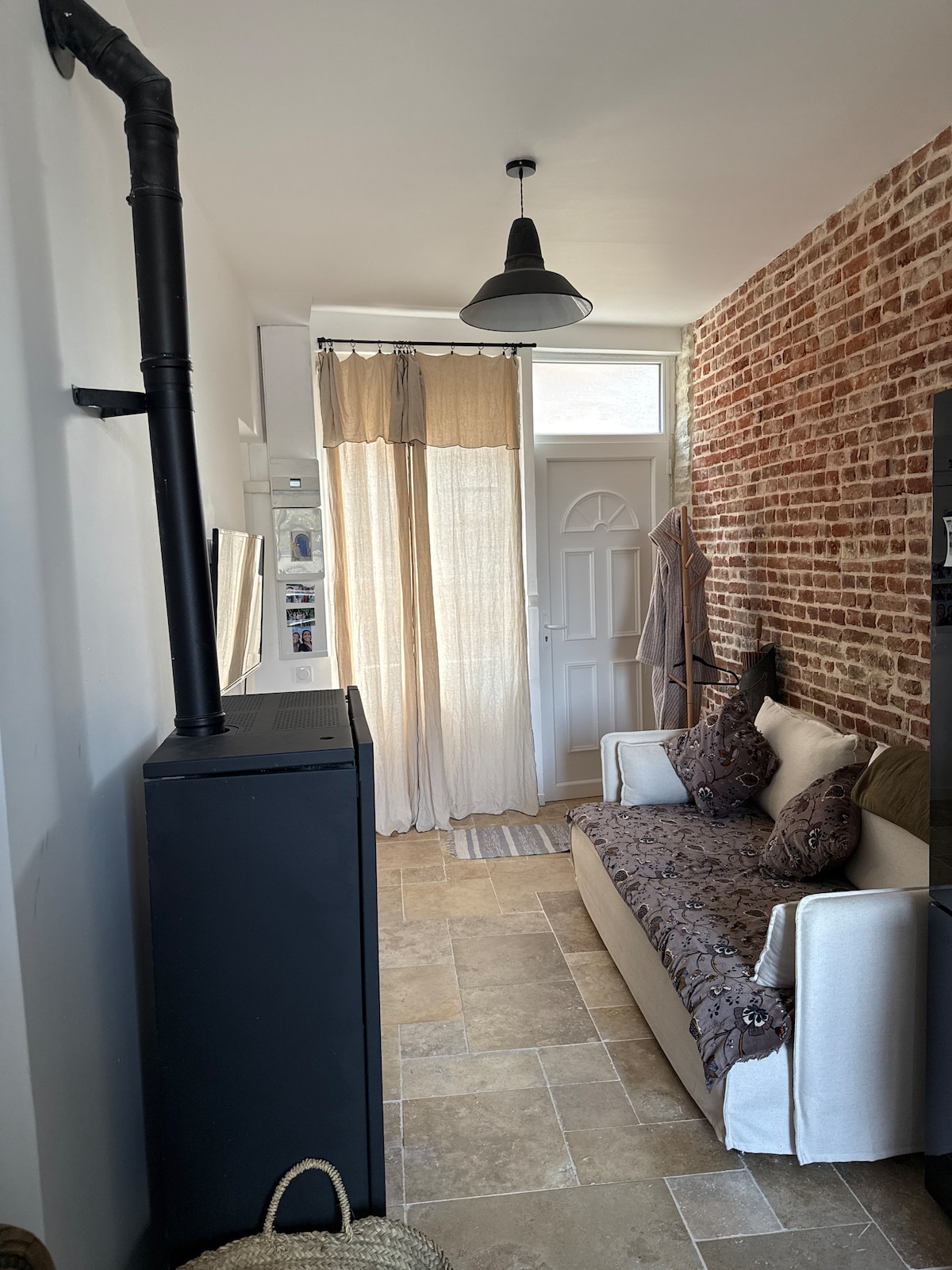 A quiet living space is showcased, featuring a cozy sofa adorned with decorative cushions. A textured brick wall and soft fabric curtains add warmth. A door allows natural light to enter, while neutral-toned tiles and a small striped rug enhance the serene ambiance.