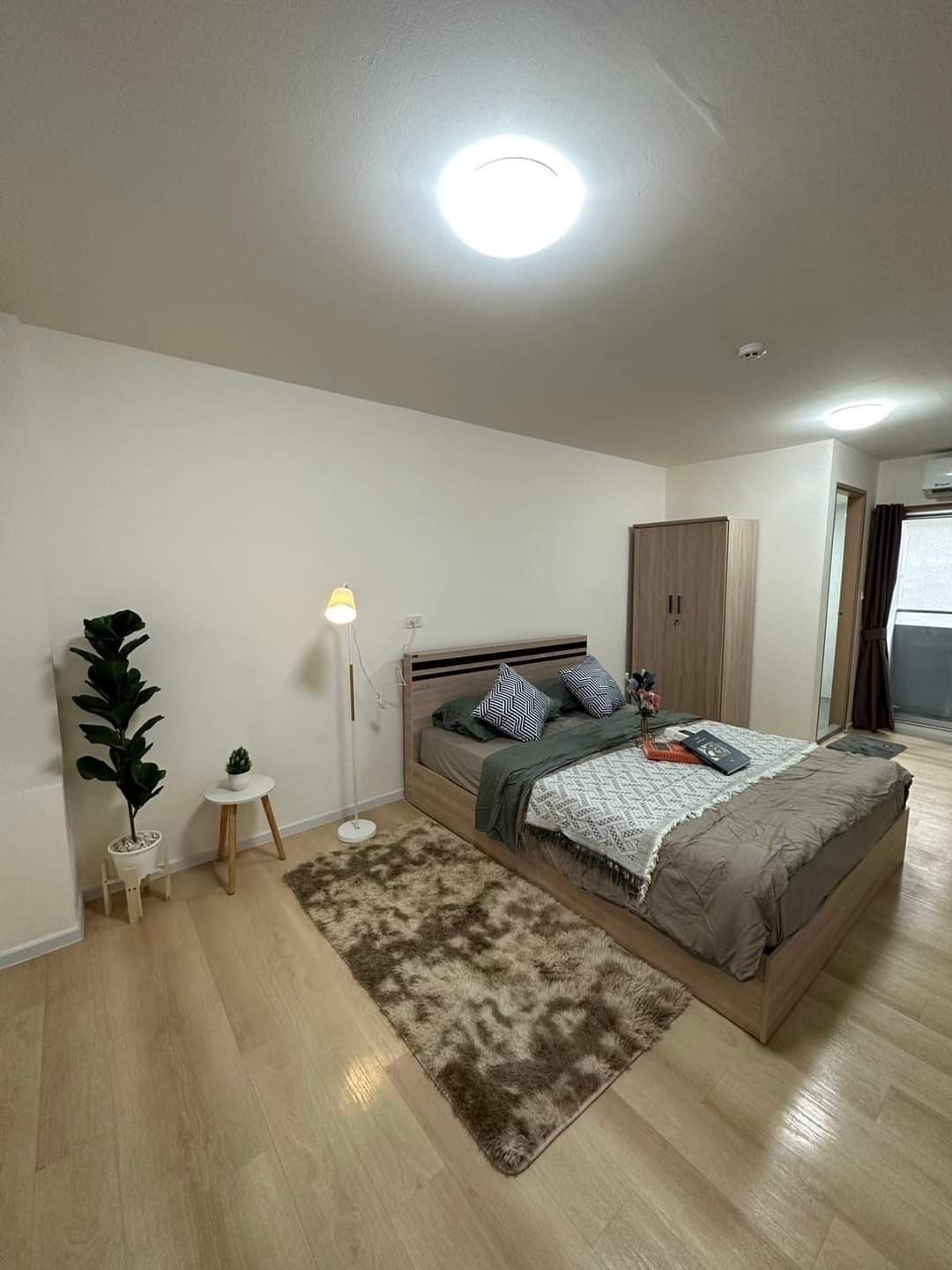 A well-lit bedroom is centered around a spacious bed, adorned with neutral-toned bedding and decorative pillows. A small side table and a lamp are positioned beside the bed. A potted plant adds a touch of greenery, while a rug provides warmth to the wooden floor.