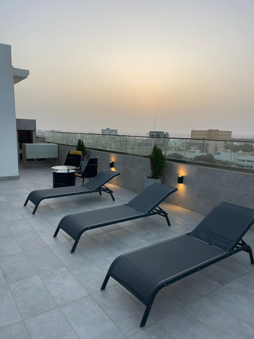 A rooftop terrace features lounge chairs arranged next to a small table. Tall planters with greenery and wall lights are positioned along the perimeter, while a soft glow from the setting sun enhances the tranquil scenery. The urban landscape is visible in the background.
