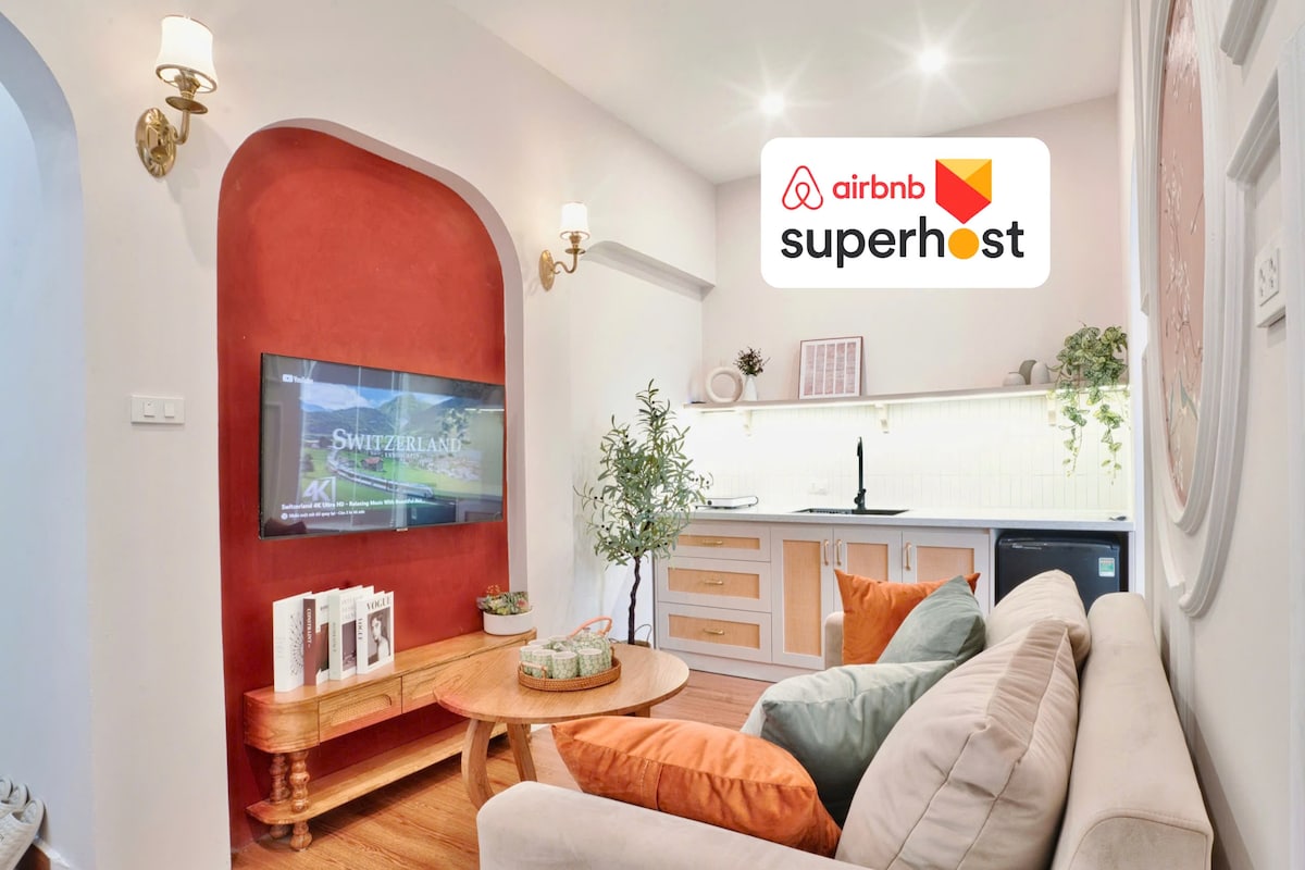 A cozy living area features a soft beige sofa complemented by earthy tones. A wooden coffee table is placed at the center, while a wall-mounted TV offers entertainment options. A small kitchen is visible in the background, showcasing modern appliances and greenery accents.