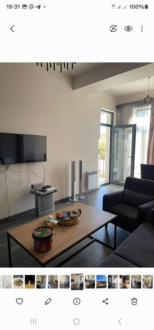 A modern living area is shown with a flat-screen television mounted on the wall. Two tall white speakers stand beside a sleek coffee table that holds a bowl of snacks. A comfortable dark couch is positioned opposite the television, and a large window lets in natural light.