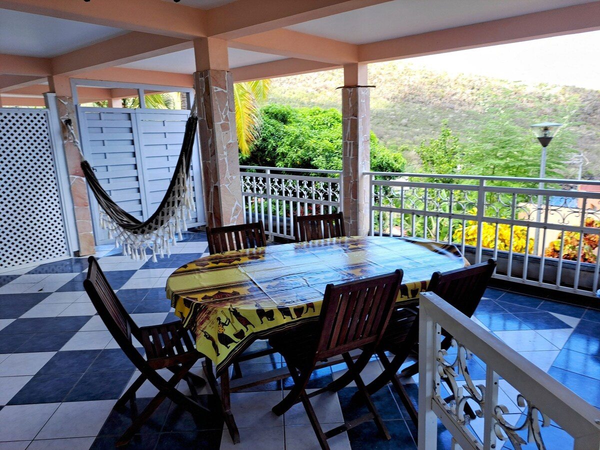 A spacious outdoor dining area features a large table surrounded by wooden chairs. A hammock is seen hanging nearby, and vibrant greenery provides a natural backdrop. The tiled floor displays a contrast of dark and light colors, enhancing the inviting outdoor space.