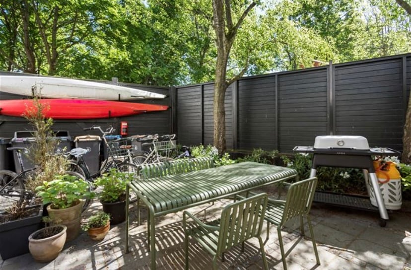 An outdoor patio area is surrounded by greenery and features a large green table with matching chairs. Two kayaks in red and white are visible above a variety of bicycles. A barbecue grill is positioned near potted plants, adding functionality to the space.