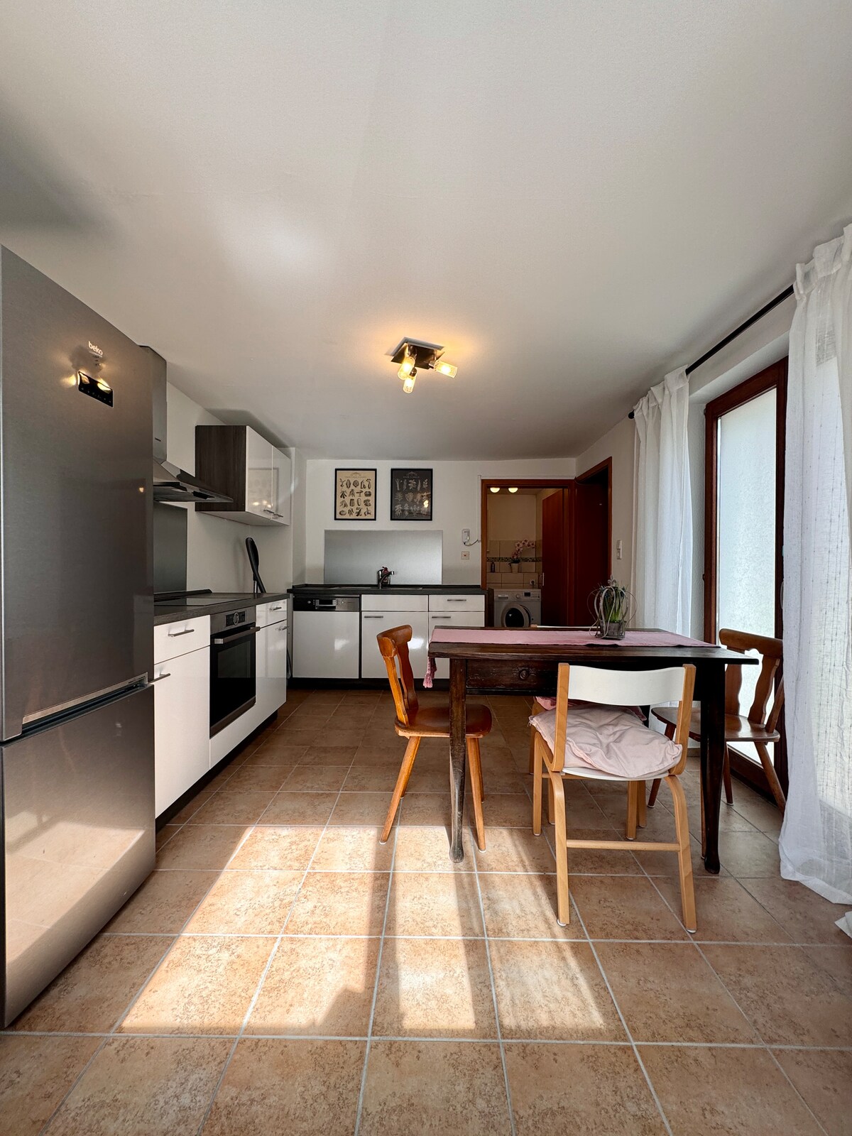 The kitchen features modern appliances, including a large refrigerator, oven, and induction cooktop. A wooden table with two chairs provides space for dining. Natural light enters through a window, complemented by sheer curtains that enhance the inviting atmosphere.