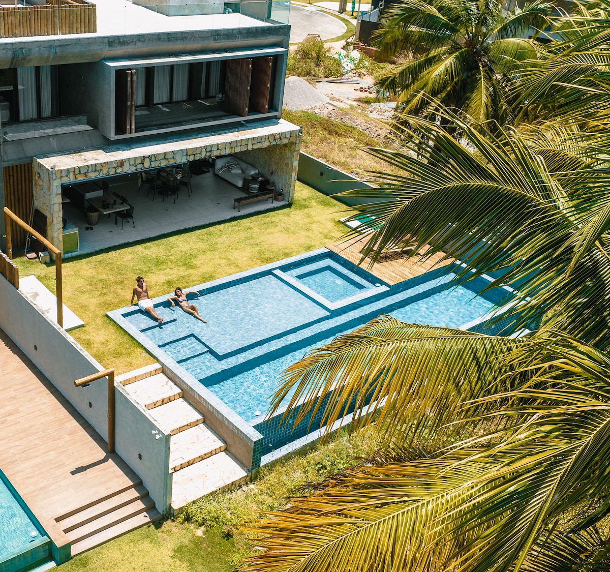 A spacious pool area is surrounded by lush green grass and palm trees, featuring two individuals relaxing in the water. The contemporary structure is partially visible, with outdoor seating available nearby.