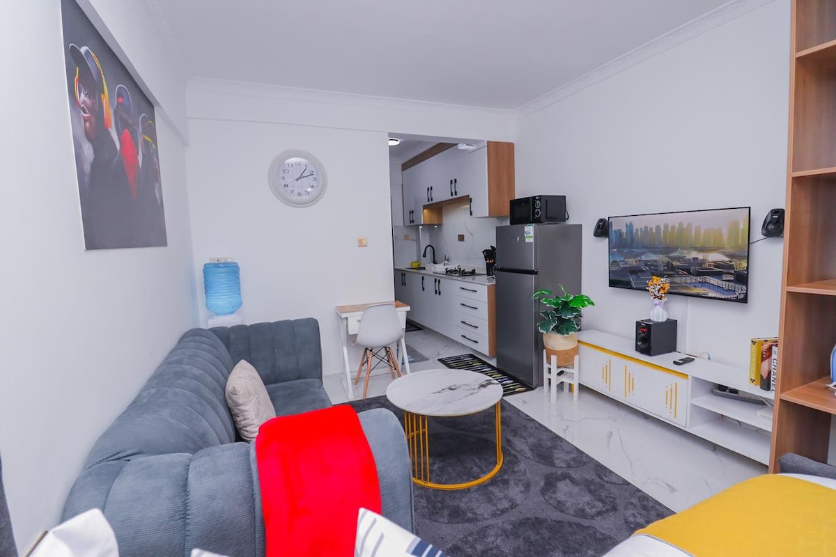 A modern living area is seen, featuring a comfortable gray sectional sofa adorned with colorful throw pillows. A round coffee table is placed in front, and a wall-mounted television is visible. The open kitchen area displays sleek cabinetry and appliances, providing a spacious feel.
