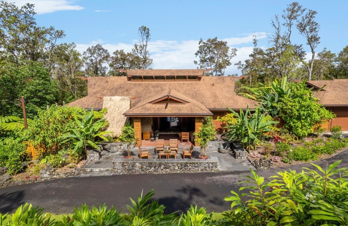 A spacious home is set among lush greenery and tall trees, featuring a welcoming entrance with a stone walkway. The covered porch showcases seating arranged in a cozy manner, inviting relaxation. The thatched roof blends harmoniously with the surrounding landscape.