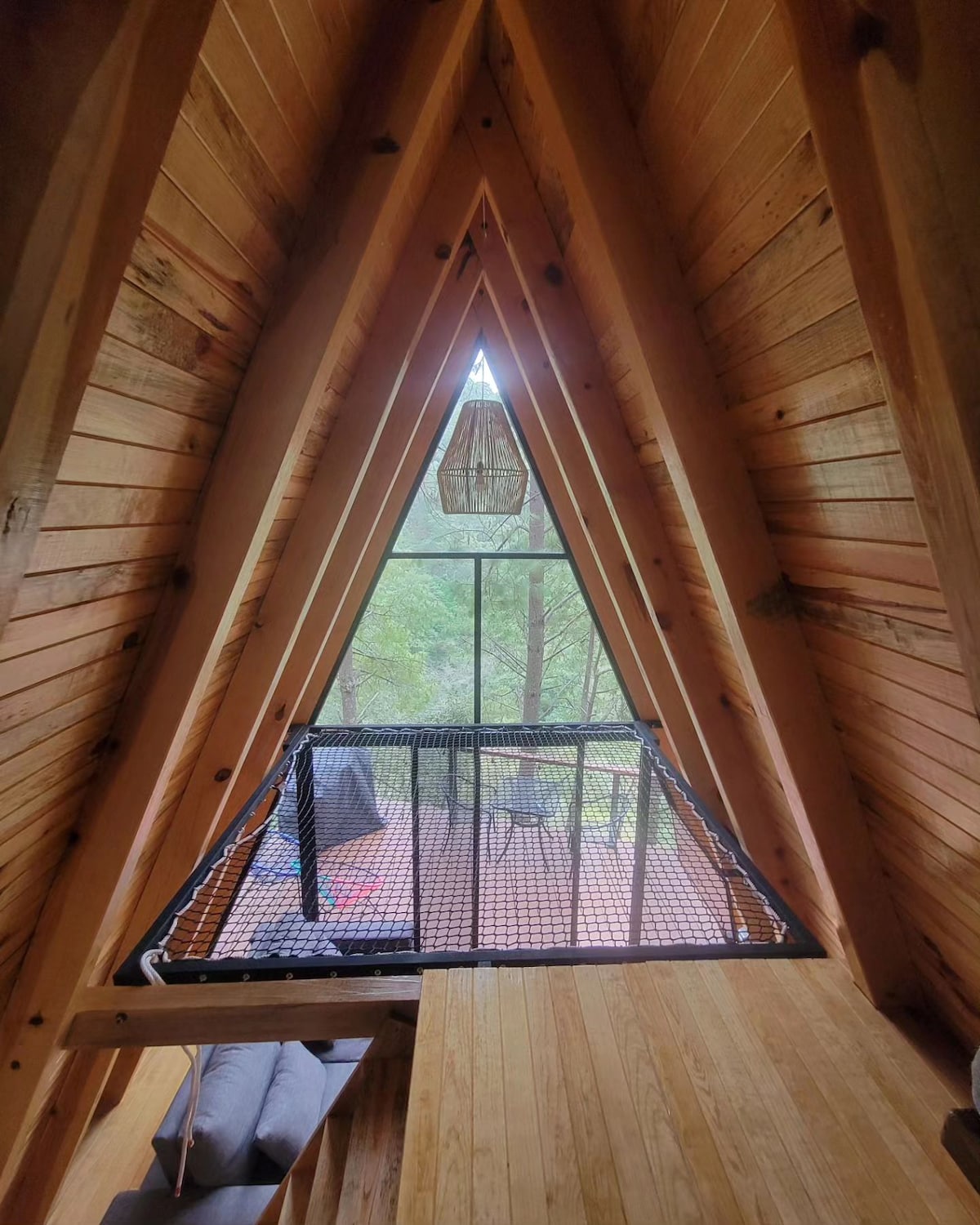 An open loft space is framed by a distinct triangular design, featuring wooden paneling and exposed beams. A netted railing overlooks a serene view of the surrounding greenery. A pendant light hangs from the peak, adding character to the space.