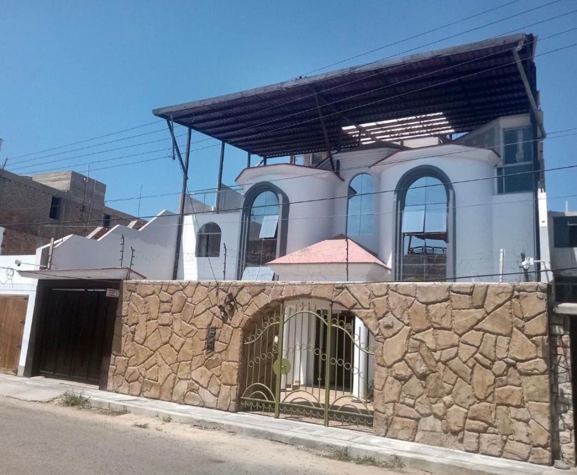 A modern two-story house features large arched windows and a distinctive roof design. The exterior is complemented by a stone wall and a gated entrance, providing a sense of privacy. Bright sunlight illuminates the facade, highlighting its architectural details.