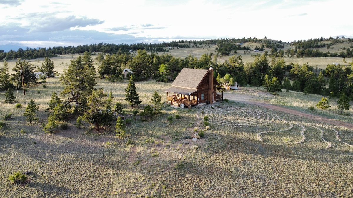 A cabin nestled in a sprawling landscape, surrounded by patches of trees and open fields. The outdoor stone meditation labyrinth is visible in the foreground, leading towards the inviting wooden structure, which features a covered deck and a charming roofline against the expansive sky.