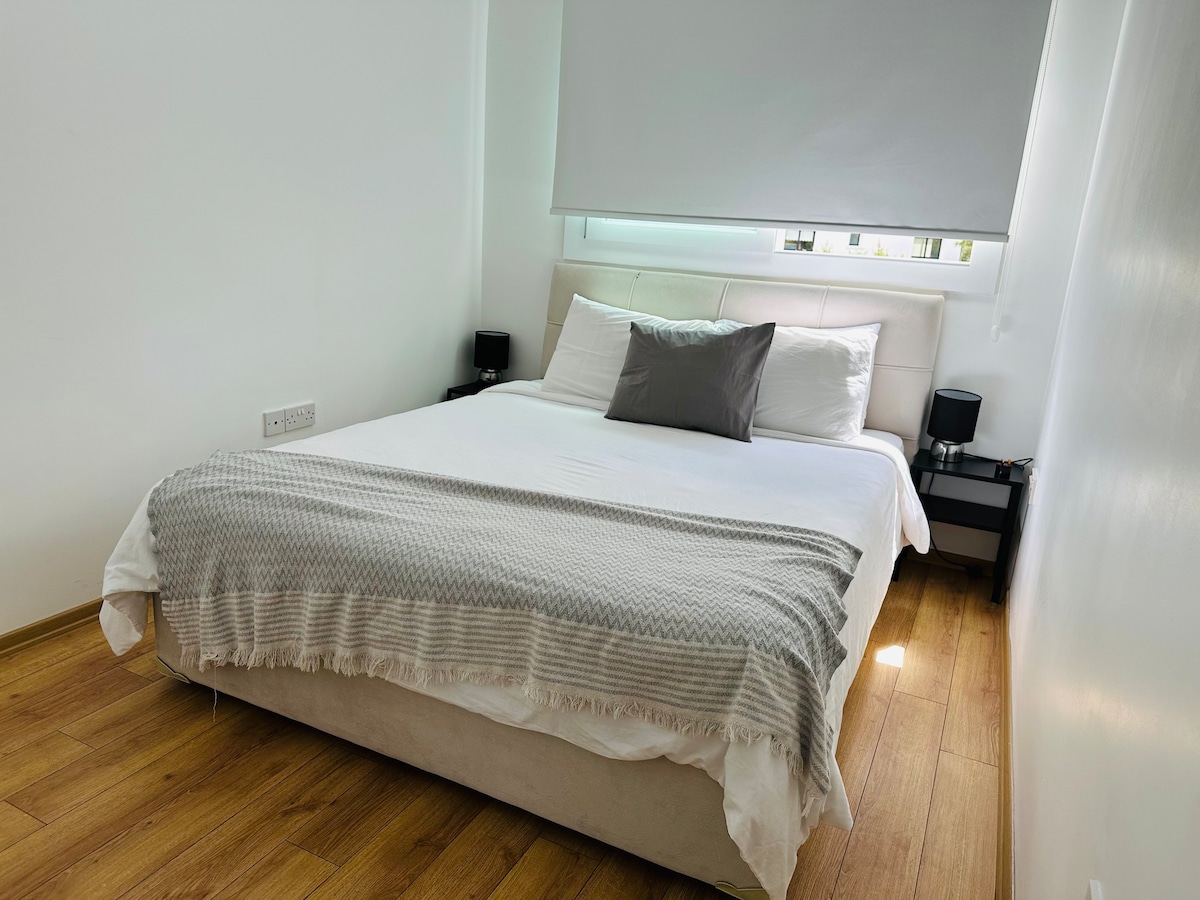 The bedroom features a spacious bed dressed in white linens with a textured gray throw. Two bedside tables are positioned on either side, each topped with a lamp. Natural light filters in through a partially covered window, illuminating the warm wooden floor.