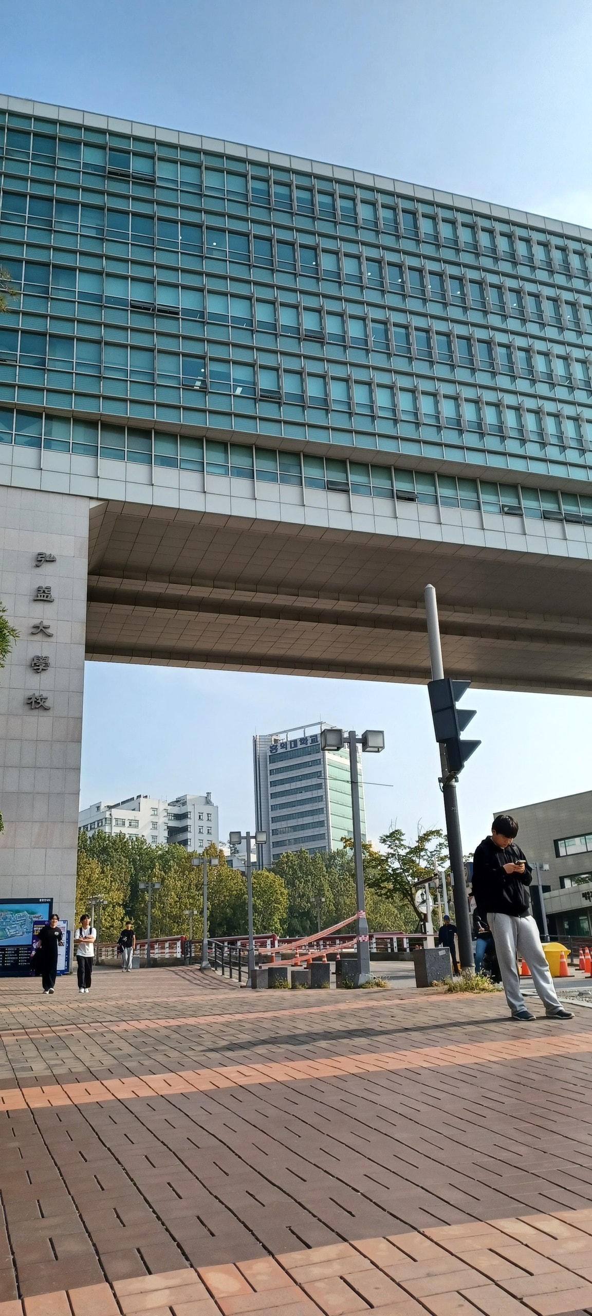 A modern building is depicted, characterized by a large glass façade that allows natural light to enter. The area features wide walkways and trees, creating a spacious environment. Pedestrians are seen walking, contributing to a lively atmosphere, with traffic lights visible for safety.